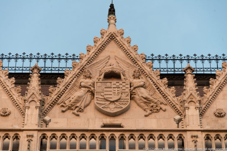 Carved Hungarian coat of arms and heraldic figures within an ornate triangular gable on the Parliament façade.