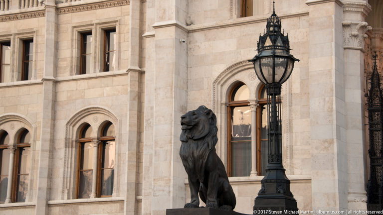 Dark lion sculpture beside a tall iron lamp and arched windows on the Hungarian Parliament exterior.