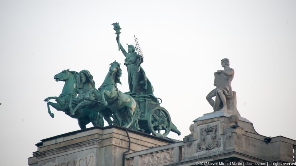 Green bronze quadriga with horses and an allegorical figure above a stone parapet and seated statue.