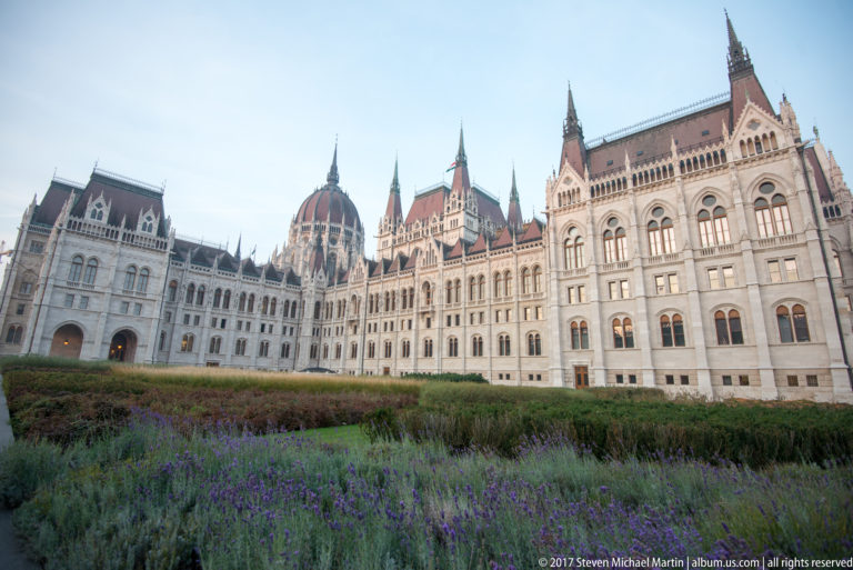 Purple flowers and garden beds in front of the Hungarian Parliament’s dome and ornate pale-stone façades.