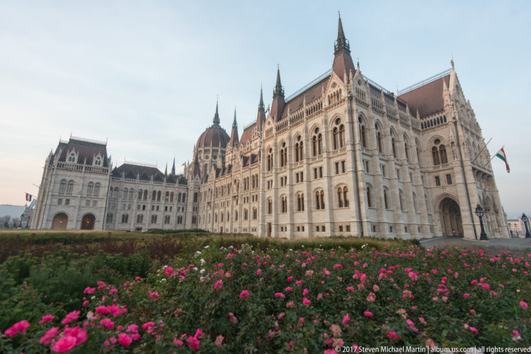 Pink flowers below a projecting Parliament pavilion, with the central dome and adjoining wings visible to the left.