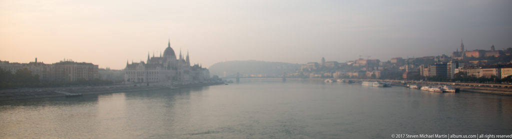 Hazy Danube cityscape with the Hungarian Parliament silhouetted on the left bank, Buda hillside at right, and a bridge in the distance.