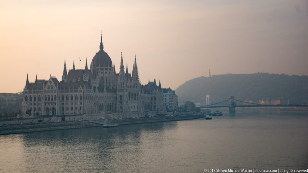 Hungarian Parliament on the Danube at left, with a downstream bridge and hazy Buda hills beyond.