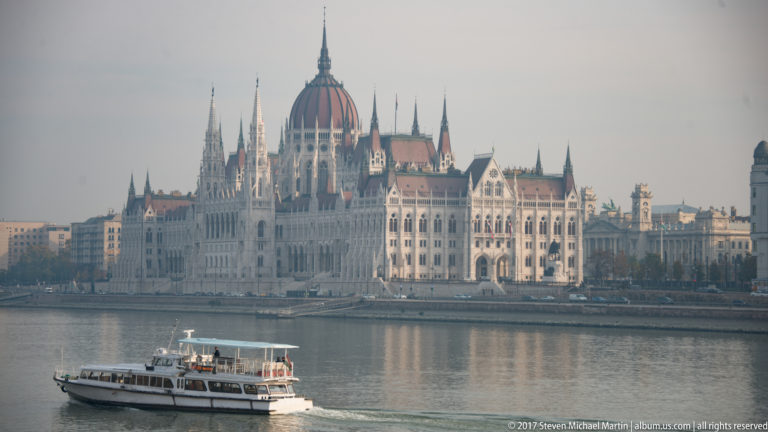 White passenger boat crossing the Danube in front of the Hungarian Parliament’s riverside façade and dome.