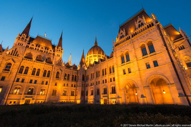 Low-angle night view along the illuminated Hungarian Parliament toward its glowing central dome.