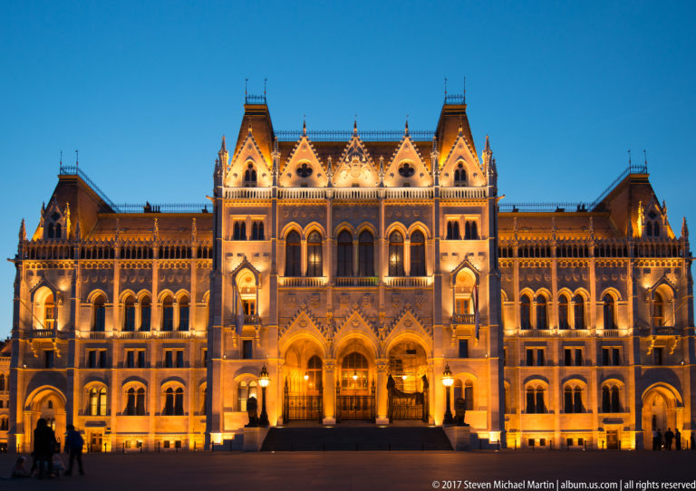 Symmetrical illuminated entrance pavilion of the Hungarian Parliament with twin arches, gables, and a rose window.