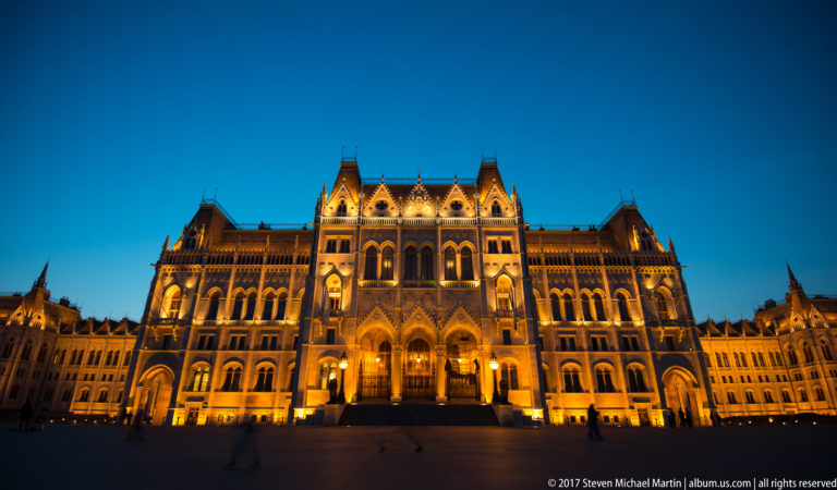 Wide blue-hour view of the illuminated Hungarian Parliament entrance across the open paving of Kossuth Lajos Square.