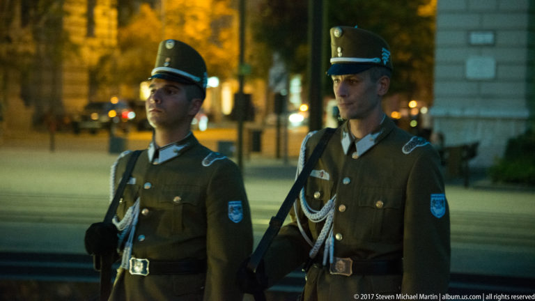 Two Hungarian ceremonial guards in peaked caps and dark dress uniforms standing together at night.