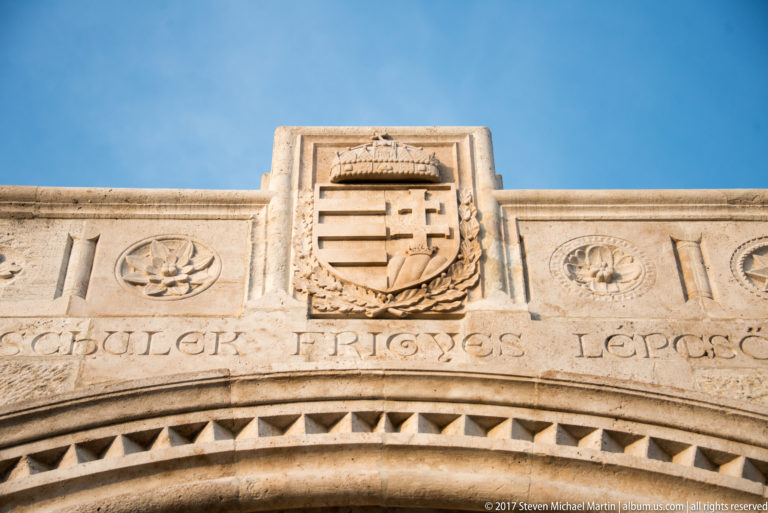 Carved Hungarian coat of arms and partially legible inscription above a stone arch at Fisherman’s Bastion.