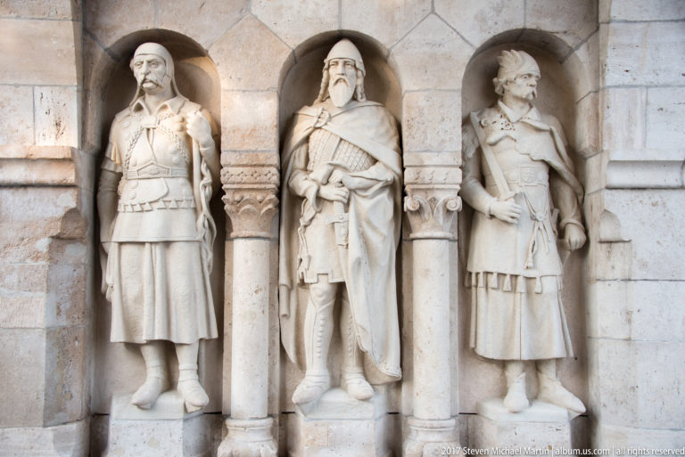 Three carved stone warriors in niches, wearing historicizing dress and carrying weapons at Fisherman’s Bastion.