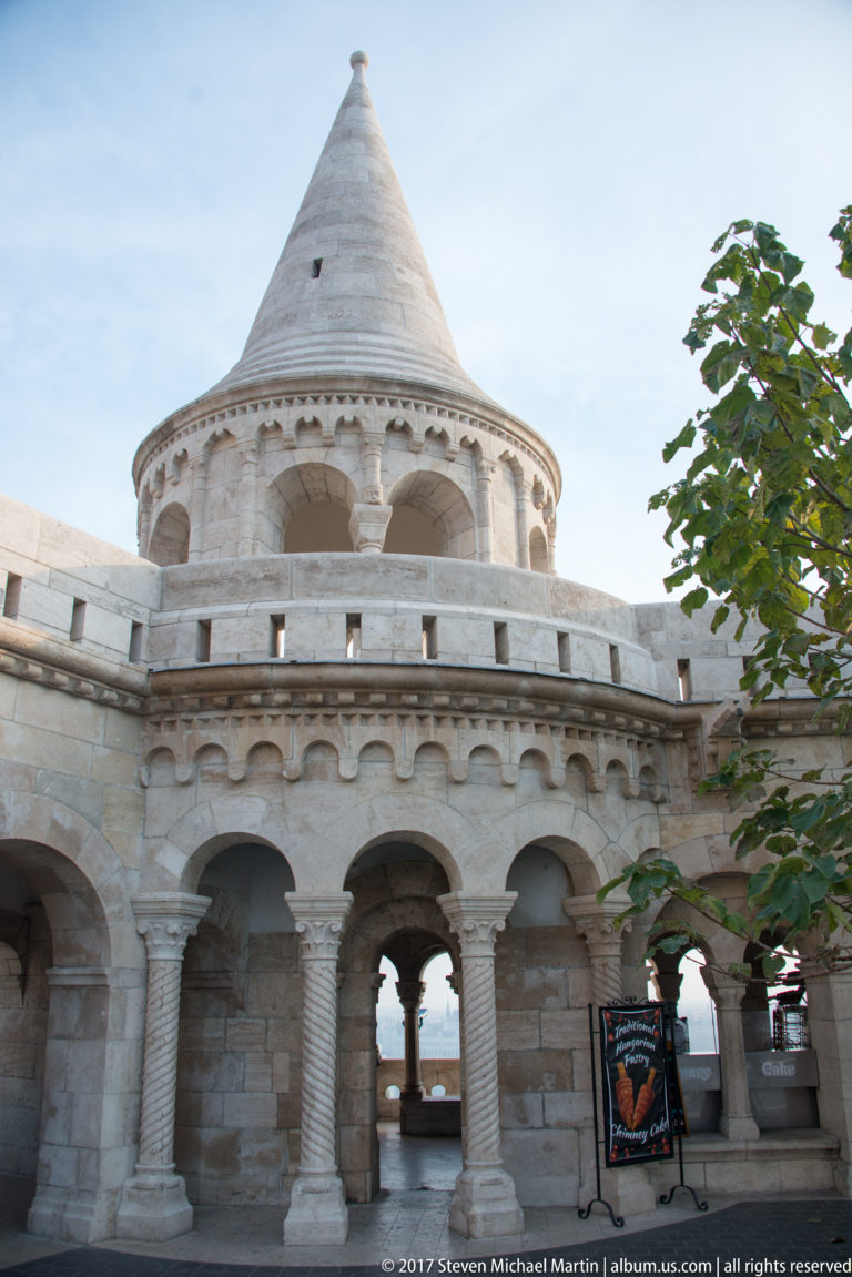 Conical turret and open round-arched colonnade of Fisherman’s Bastion, seen from below beside leafy branches.