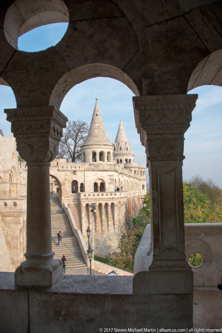 Fisherman’s Bastion terraces and conical towers framed through a shaded stone arcade and thick columns.