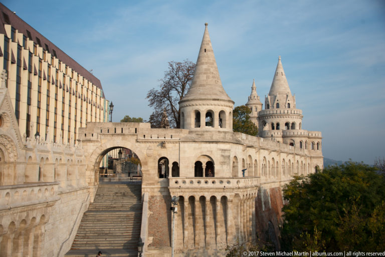 Broad stairway and arched gateway between conical turrets and arcades at Fisherman’s Bastion, beside a modern building.