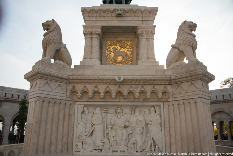 Carved pedestal of the Saint Stephen I monument with historical relief, gilded medallion, and flanking stone lions.