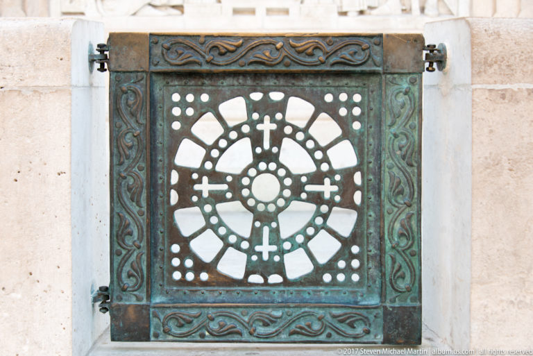Patinated square bronze grille with circular pierced pattern and small crosses set into pale stone at Fisherman’s Bastion.