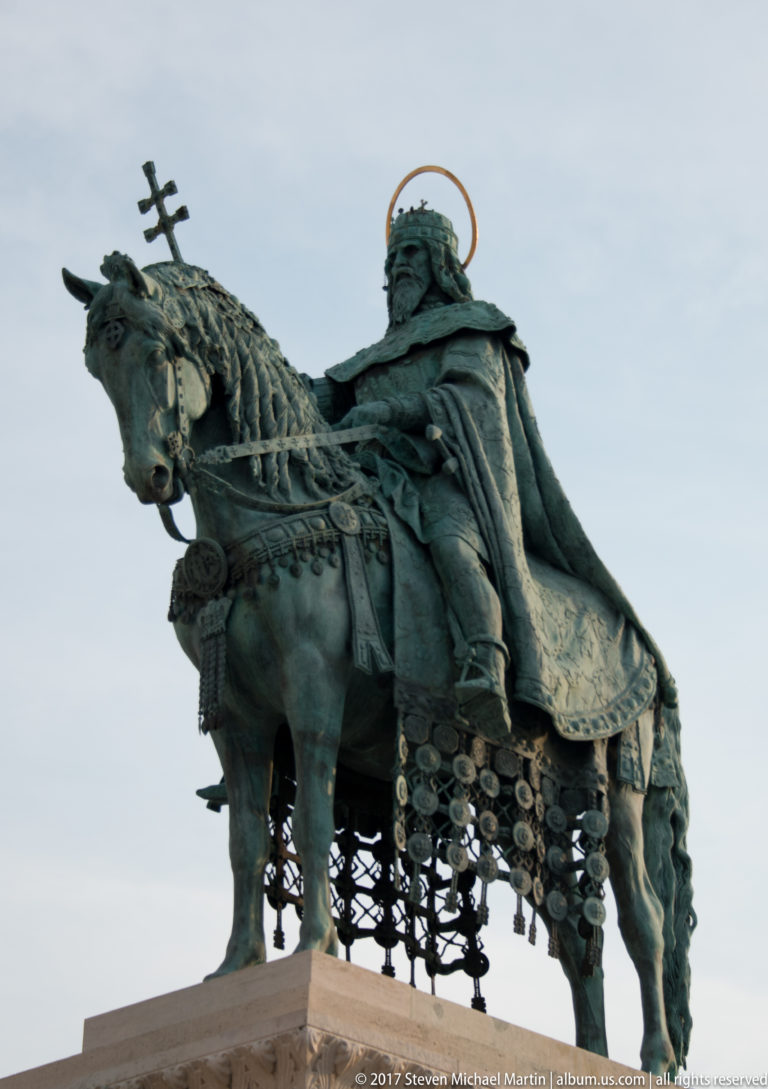 Close side view of the bronze Saint Stephen I equestrian statue, showing crown, cloak, horse tack, and gilded halo-like ring.