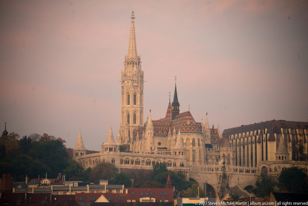 Distant daylight view of Matthias Church, its patterned roof and spire rising above Fisherman’s Bastion and Buda rooftops.
