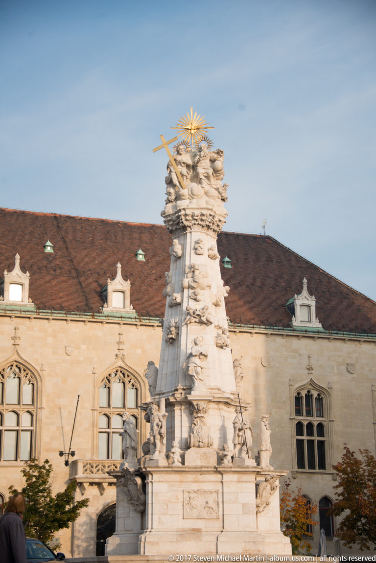 Tall sculpted Holy Trinity Column in Budapest’s Castle District, topped by clustered figures and gilded rays before a historic building.