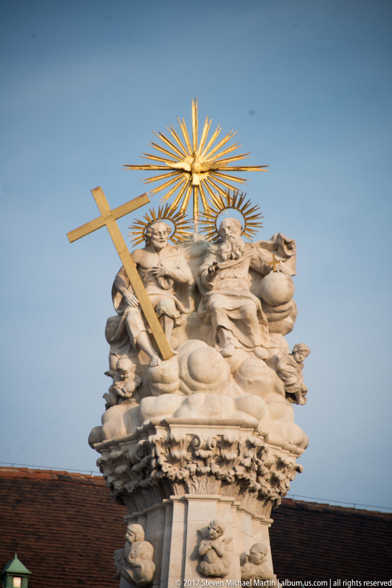 Close view of the Holy Trinity Column’s sculpted crown with clustered figures, a large cross, and gilded radiating rays.