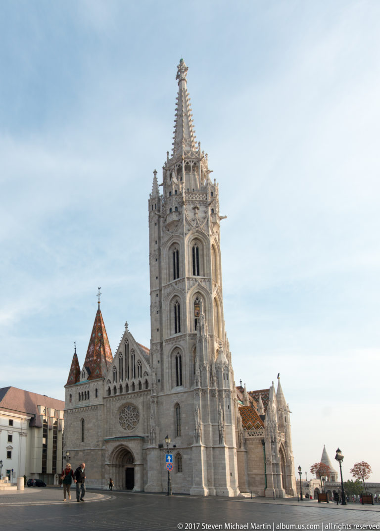 Frontal view of Matthias Church with pointed entrance, rose window, patterned roof, and tall Gothic Revival spire from the square.