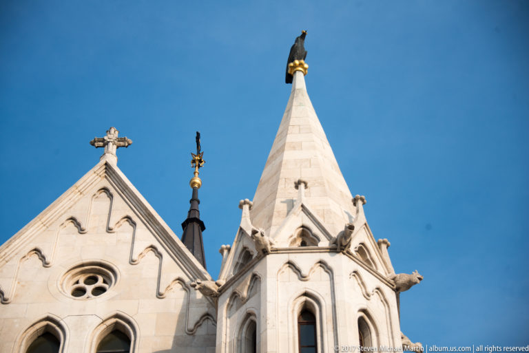Pale stone gable and pinnacled turret of Matthias Church with crosses, pointed openings, and projecting carved spouts against blue sky.