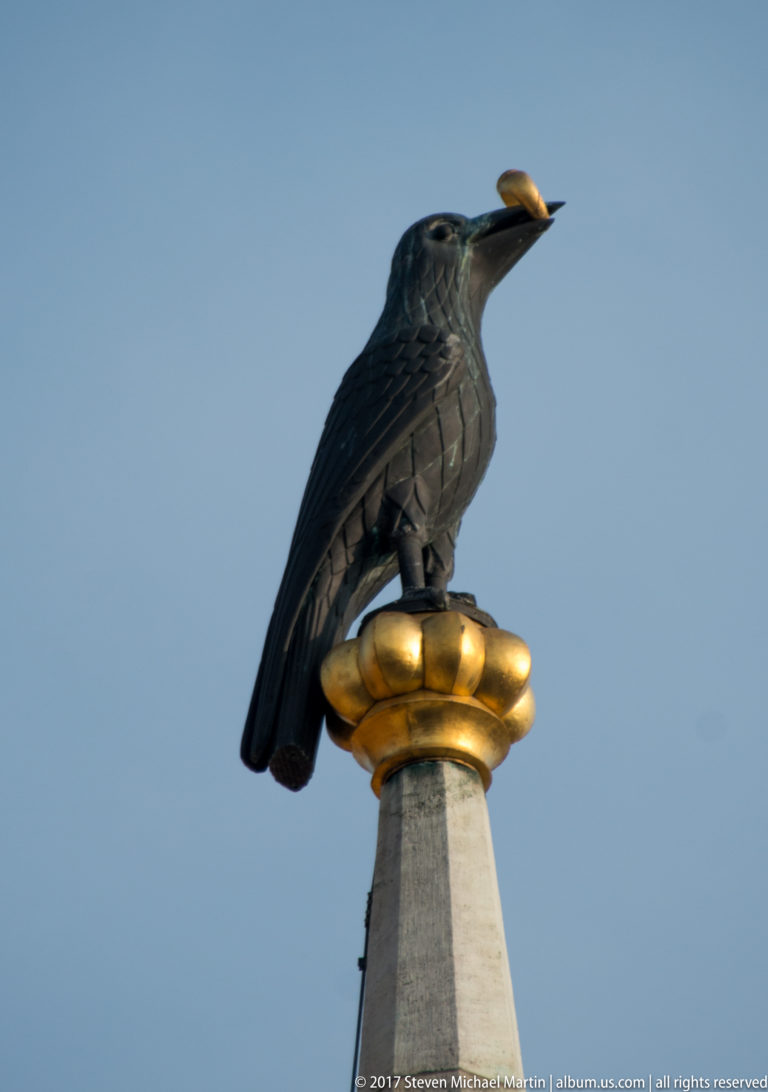 Dark raven-like bird sculpture perched on a gilded architectural finial against a clear blue sky.