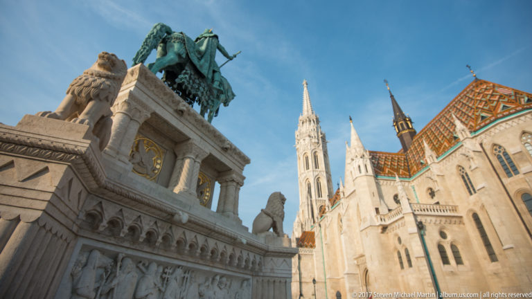 Low-angle view of Matthias Church with patterned tile roof, pale stone balustrades, sculptural ornament, and a bronze equestrian monument.