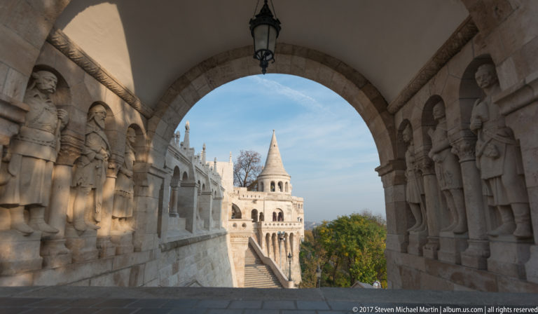 Stone arcade lined with sculpted figures, framing stairs and a conical-roofed tower at Fisherman’s Bastion.