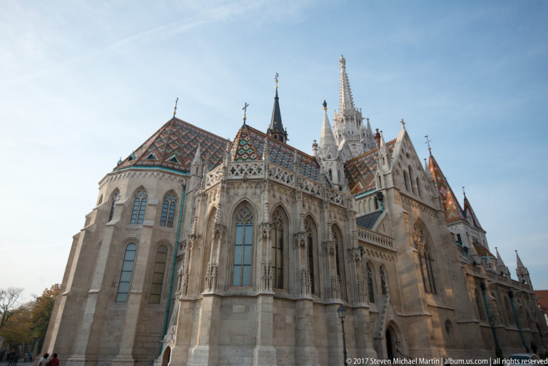 Exterior of Matthias Church showing buttressed apse, tall Gothic windows, spires, and colorful patterned roof tiles.