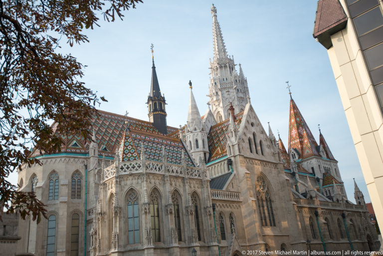 Matthias Church exterior with tall Gothic windows, buttresses, pinnacles, and multicolored patterned roof behind autumn foliage.