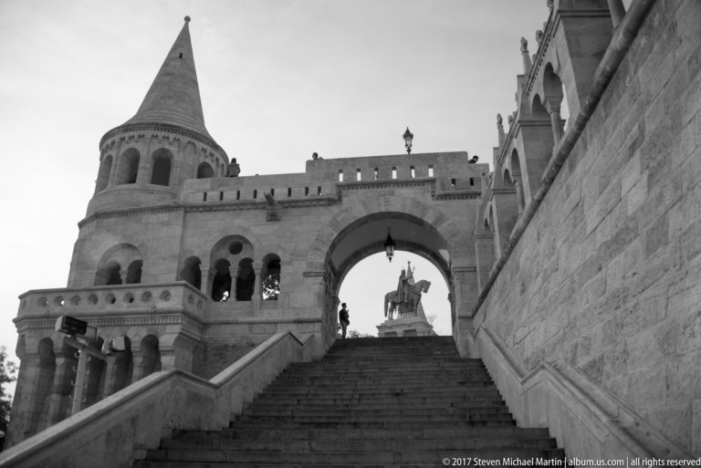 Black-and-white view up stone stairs beneath a Fisherman’s Bastion arch, with conical tower and equestrian statue beyond.