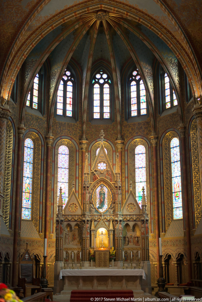 Symmetrical Matthias Church sanctuary with altar, tall stained-glass windows, ribbed vaulting, and richly painted interior surfaces.