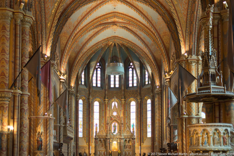Long interior view of Matthias Church toward the sanctuary, with painted arches, stained glass, and carved pulpit at right.