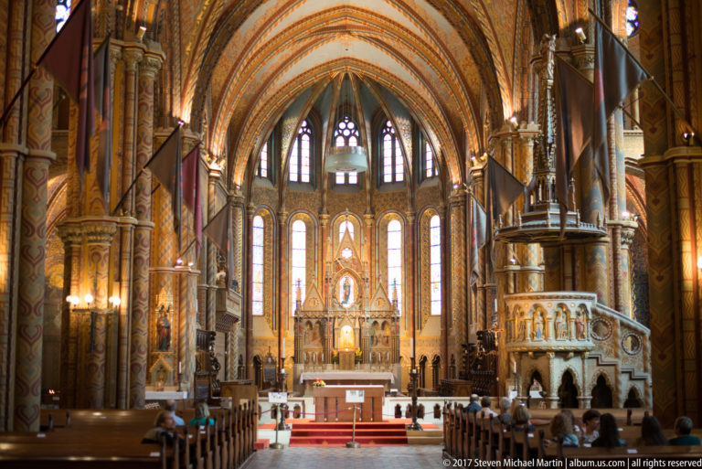 Wide Matthias Church interior with pews, visitors, carved pulpit, altar, stained glass, and painted ribbed vaulting.