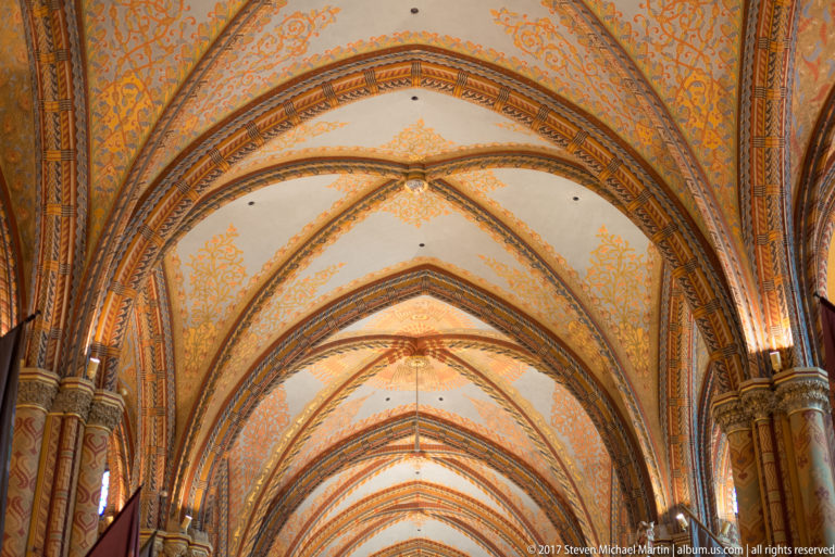 Upward view of Matthias Church rib vaults with pale panels, orange foliate ornament, and dark patterned ribs.