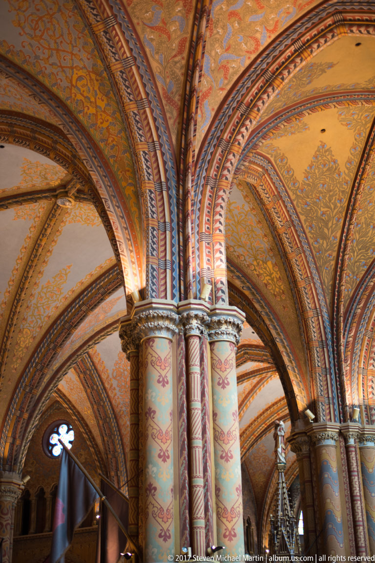 Painted clustered columns rising into intersecting ribbed arches inside Matthias Church, all covered with polychrome ornament.
