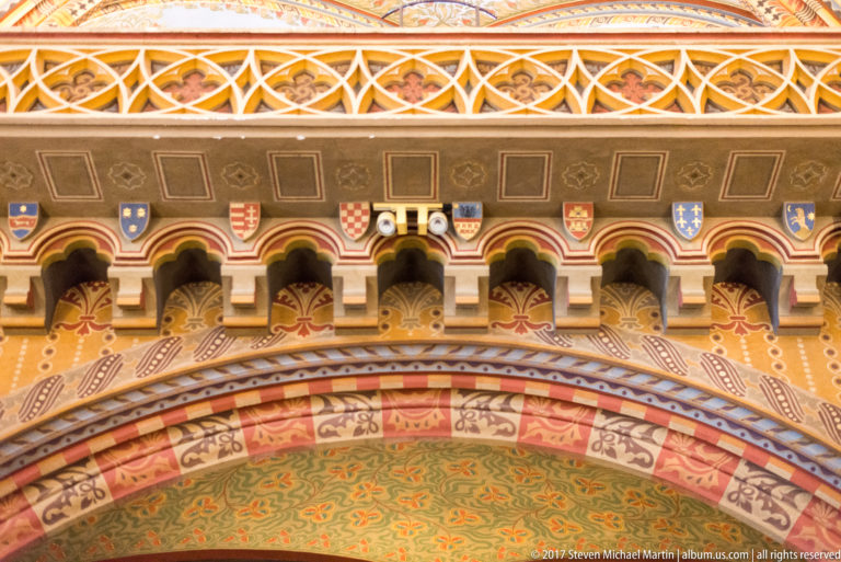 Heraldic shields above a small arched gallery, surrounded by red, green, gold, and cream painted ornament.