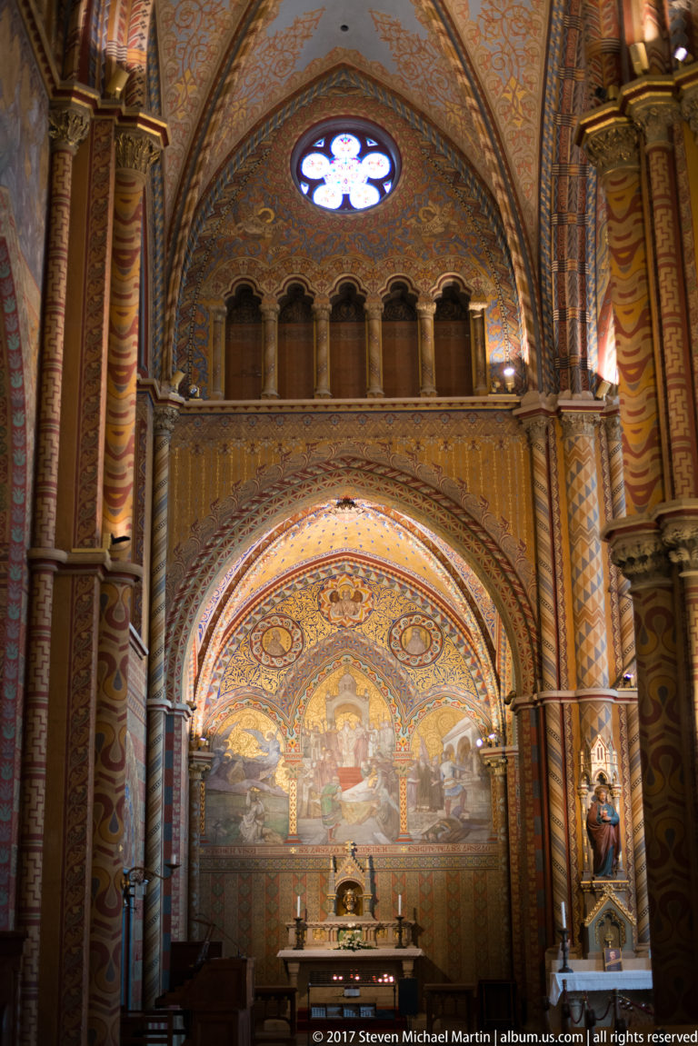 Decorated Matthias Church side chapel with painted arch, circular window, wooden gallery, murals, and small altar below.