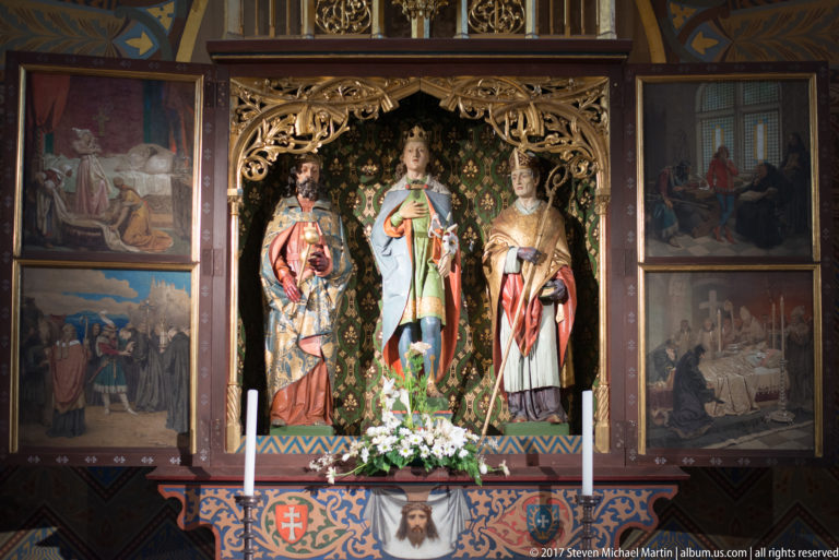 Polychrome church shrine with three standing sacred figures, carved tracery, flanking painted panels, flowers, and candles.