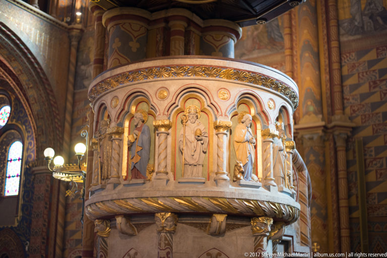 Rounded carved pulpit with standing saint figures in arched niches inside the richly decorated Matthias Church interior.
