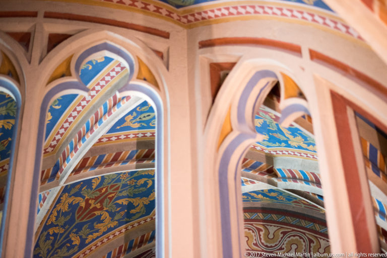 Close view through painted Gothic tracery, with blue heraldic fields and multicolored patterned decoration beyond.