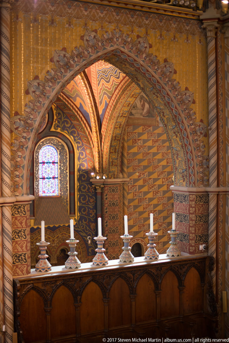 Small wooden altar with white candles beneath a painted pointed arch and stained-glass window inside Matthias Church.