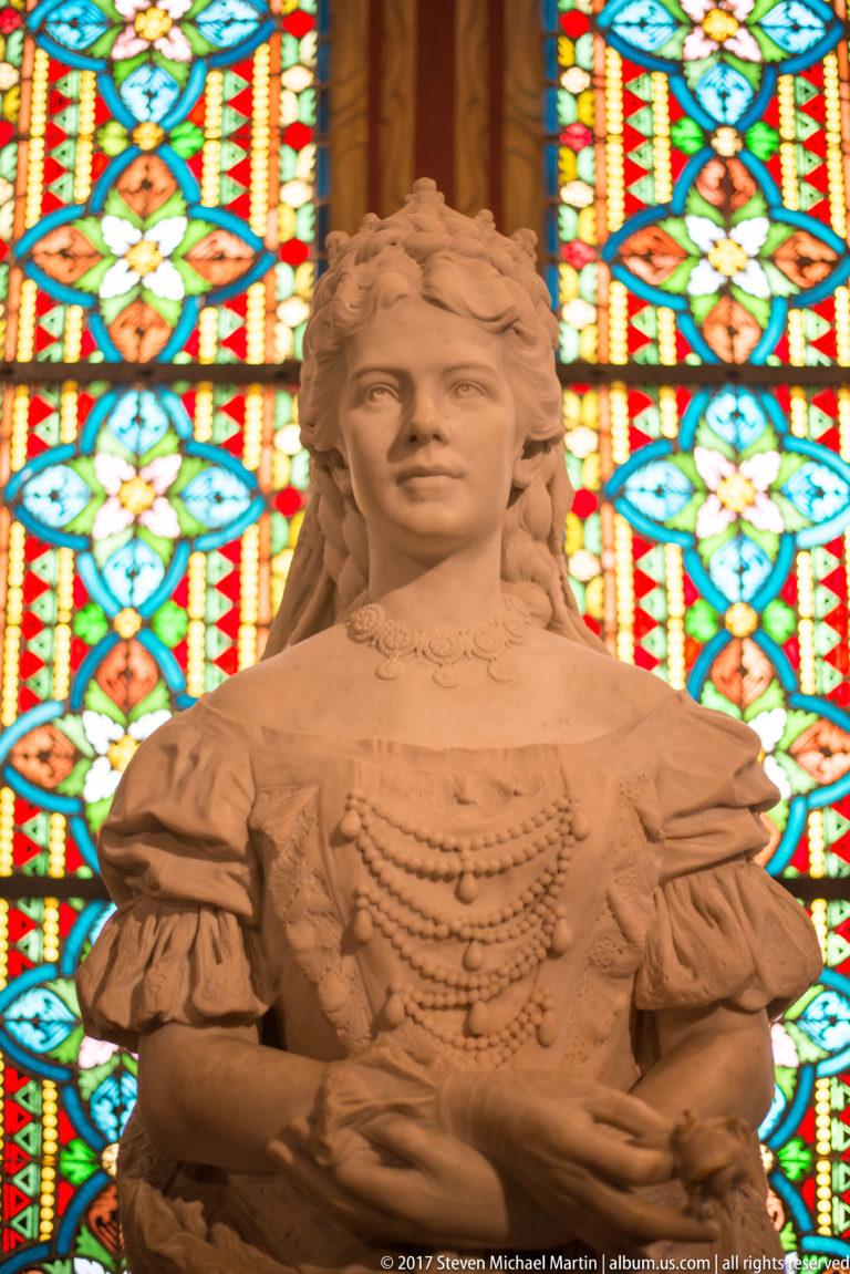 Warm-toned sculpture of a young woman in historical dress standing before a brightly colored stained-glass window.