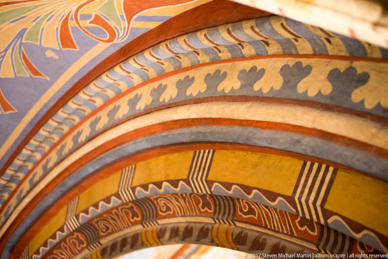 Close detail of a church arch painted with yellow, gray, red, cream, and black geometric and leaf-like bands.