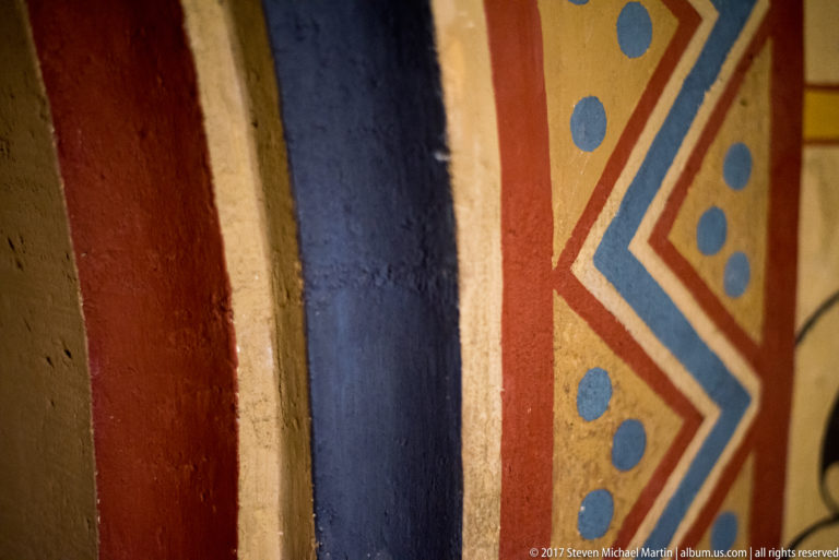 Close-up of vertical red, blue, cream, and ochre painted bands beside a zigzag border with blue dots.