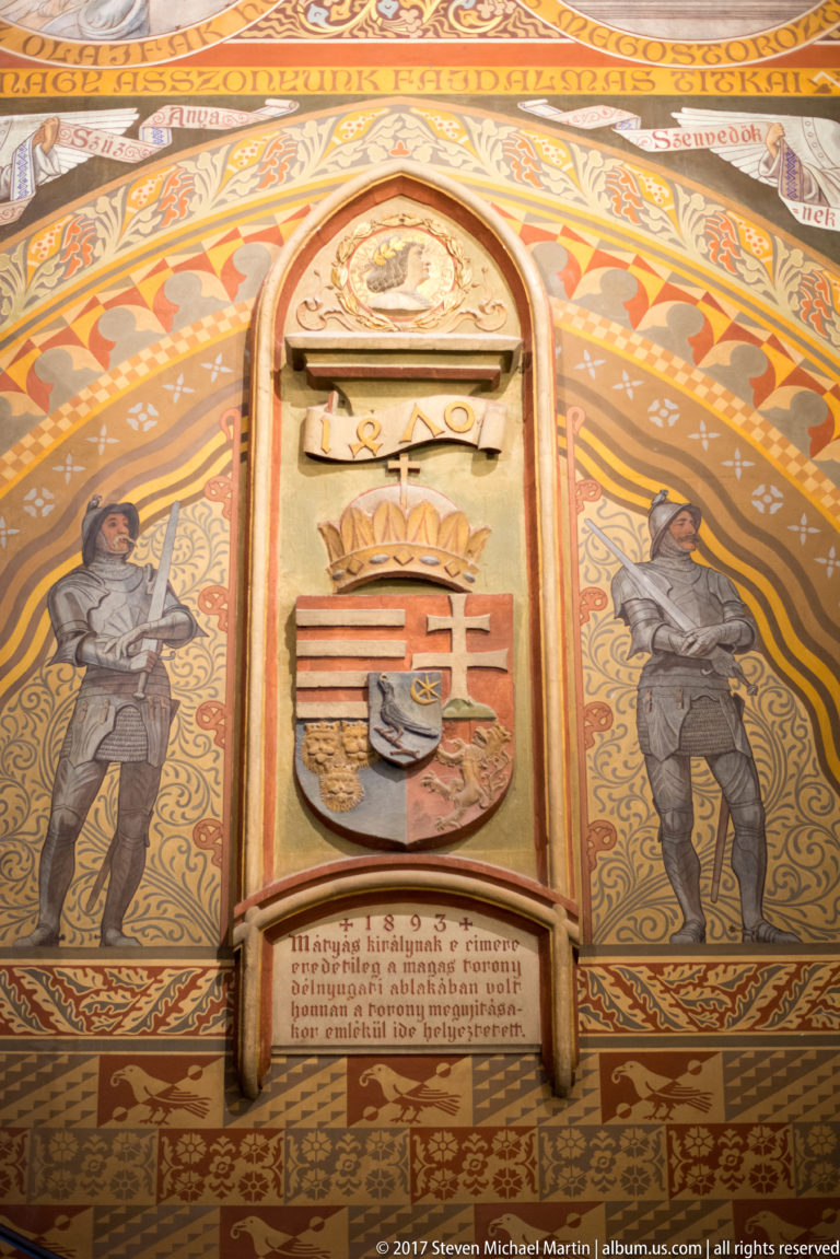 Crowned Hungarian coat of arms centered between two painted warrior figures on a richly patterned wall inside Matthias Church.