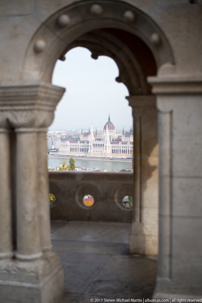 Hungarian Parliament Building across the Danube framed by a shaded stone arch at Fisherman’s Bastion.