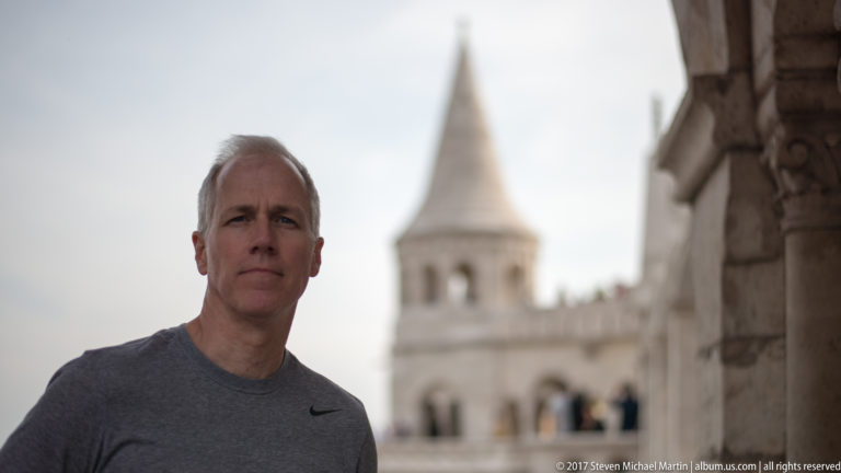 Adult man facing the camera with a blurred conical tower and arcades of Fisherman’s Bastion behind him.