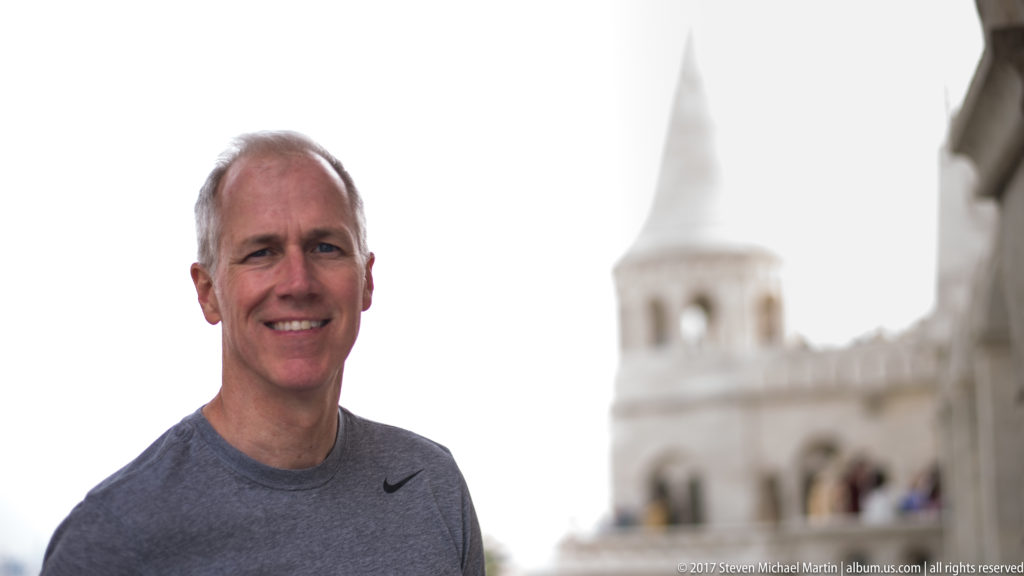Smiling adult man in the foreground with a softly blurred Fisherman’s Bastion tower and arcades behind him.