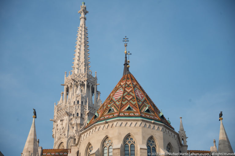 Close view of Matthias Church’s patterned tile roof and tall stone spire against a clear blue sky.